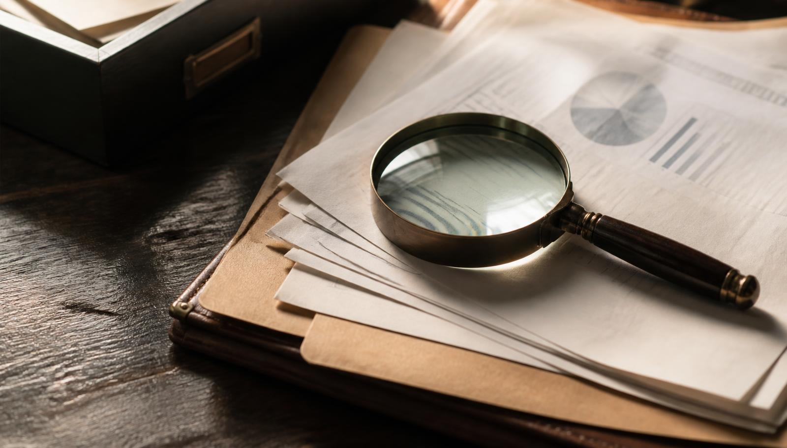 Symbolbild Due Diligence: Lupe auf gestapelten Unternehmensunterlagen und Aktenmappe